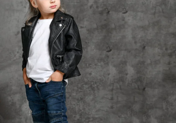 Klein schattig glimlachend blond meisje in stijlvolle rock stijl zwart lederen jas en blauwe jeans staan over grijs beton achtergrond — Stockfoto