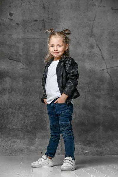 Little smiling blond girl in stylish rock style black leather jacket, blue jeans and white sneakers standing with hands in pockets Royalty Free Stock Photos