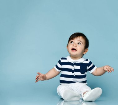 Çizgili tişörtlü, beyaz pantolonlu ve popolu küçük çocuk. Yukarı bakıyor, mavi arka planda yerde oturuyor. Kapat.