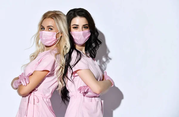 Pembe önlüklü, tek kullanımlık eldivenli ve beyaz üzerine izole edilmiş tıbbi maskeli iki kadın. Tıbbi personel, kozmetik uzmanı. Kapat.