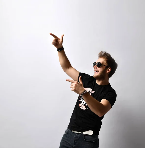 Hipster companheiro de óculos de sol, t-shirt preta com estampa, pulseiras. Ele está apontando para alguém, posando isolado no branco. Fechar — Fotografia de Stock