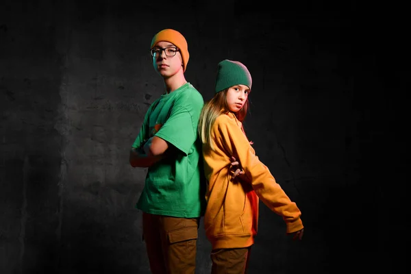 Adolescentes jóvenes niño y niña en verde y amarillo elegante ropa casual y sombreros de pie espalda con espalda sobre fondo oscuro — Foto de Stock