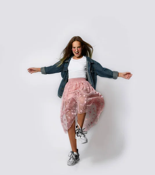 Young positive teen girl in stylish casual clothes and sneakers standing and moving with hands stretched out over white background — Stockfoto