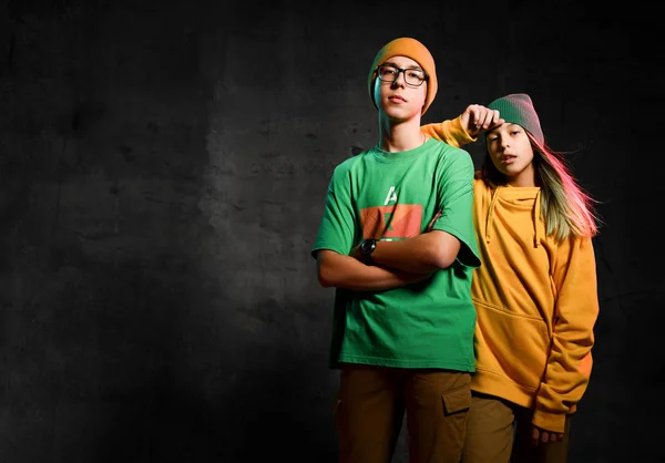 Adolescentes jóvenes niño y niña en verde y amarillo elegante ropa casual y sombreros de pie y mirando a la cámara sobre el fondo oscuro — Foto de Stock