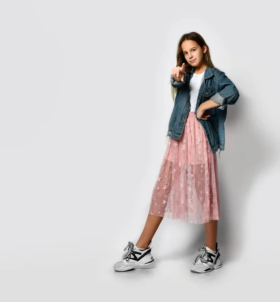 Young smiling teen girl in stylish casual clothes and sneakers standing and pointing at camera with finger over white background — ストック写真