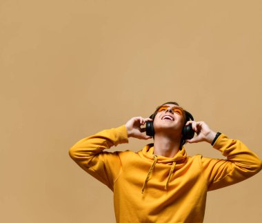Young positive teen boy in yellow hoodie, sunglasses and headphones standing, looking up and smiling over yellow wall background