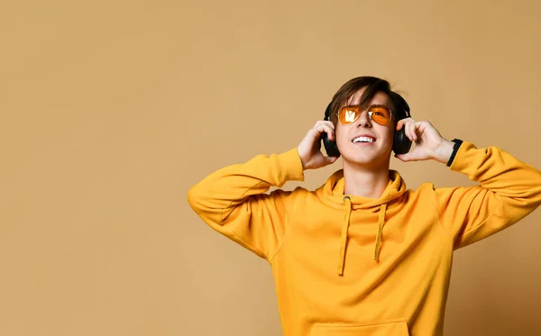 Joven adolescente positivo con capucha amarilla, gafas de sol y auriculares de pie y sonriendo —  Fotos de Stock