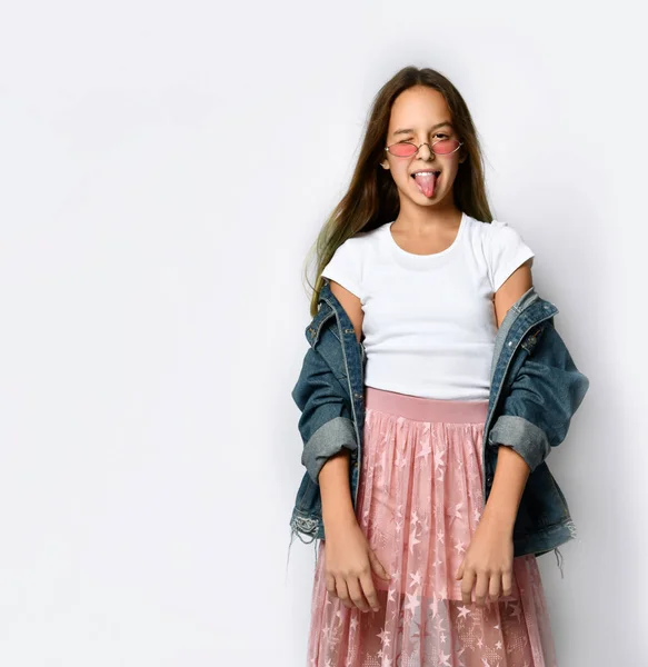 Молода дівчина-підліток у стильному повсякденному одязі та рожевих сонцезахисних окулярах стоїть, відчуває себе грайливим та показує язик на білому тлі — стокове фото