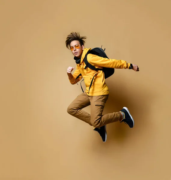 Young teen boy in comfortable clothing, sneakers and backpack jumping over yellow wall background — Φωτογραφία Αρχείου