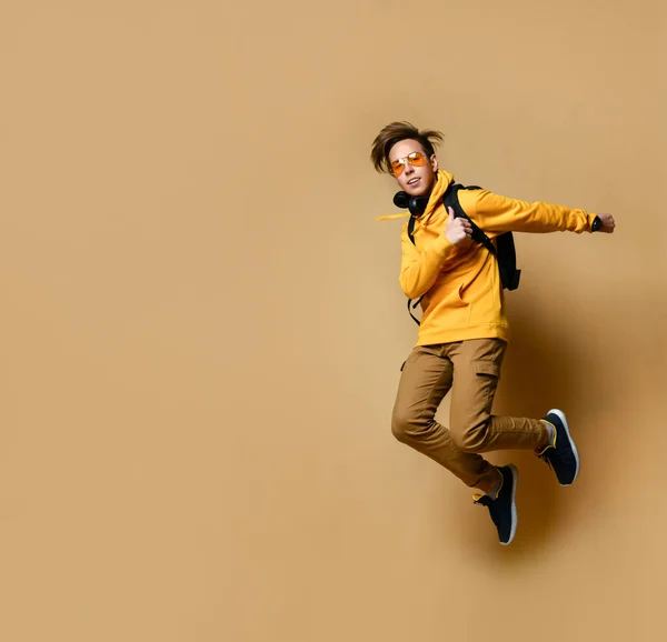 Young teen boy in comfortable clothing, sneakers, sunglasses and backpack jumping and feeling cool over yellow wall background — Φωτογραφία Αρχείου