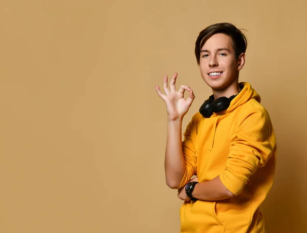 Young positive teen boy in yellow hoodie and headphones standing, smiling and showing good luck gesture over yellow background — 스톡 사진