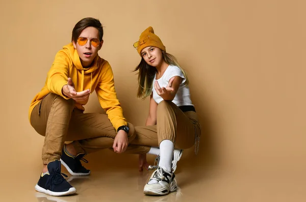 Young teens boy and girl in comfortable clothing, hats, sneakers and sunglasses sitting and pointing at camera — Φωτογραφία Αρχείου