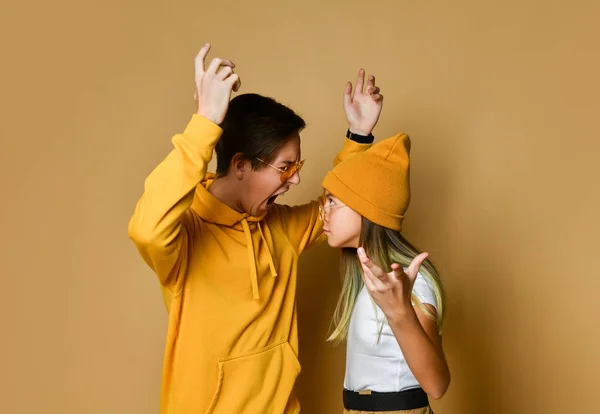 Young teens boy and girl in comfortable clothing, hats and sunglasses standing and feeling angry over yellow background — ストック写真