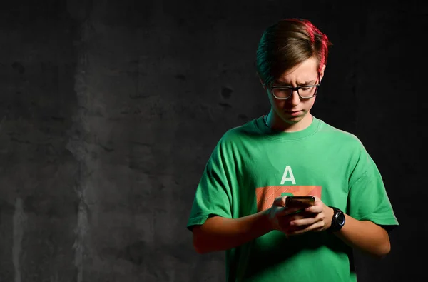 Young positive teen boy in green t-shirt and glasses standing, texting message on phone and smiling over dark background — Stockfoto