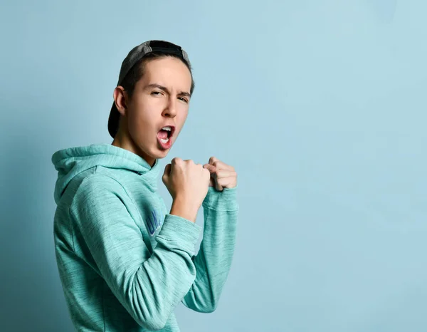 Young teen boy in blue hoodie standing, shouting and ready to defend himself with fists over pastel blue wall background — 스톡 사진