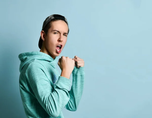 Joven adolescente con capucha azul de pie, gritando y listo para defenderse con los puños sobre fondo de pared azul pastel —  Fotos de Stock