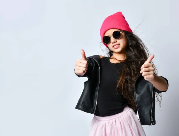 Teenage female in black leather jacket, pink hat and sunglasses. She smiling, showing thumbs up, isolated on white. Close up — 图库照片