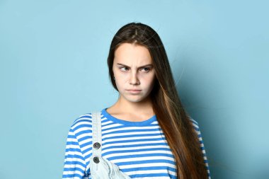 Kot pantolonlu, çizgili tişörtlü bir ergen. Alınmışa benziyor. Mavi arka planda katlanmış ellerle poz veriyor. Kapat.