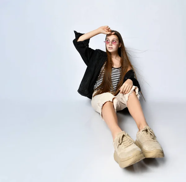 Adolescente de t-shirt listrada, casaco de ganga preta, calças e tênis. Ela está sentada no chão isolada em branco. Fechar — Fotografia de Stock