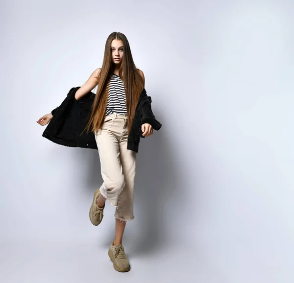 Mujer adolescente con camiseta a rayas, chaqueta vaquera negra, pantalones beige y zapatillas de deporte. Te mira, posando aislada sobre blanco. De cerca. —  Fotos de Stock