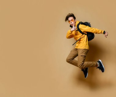 Young teen boy in comfortable clothing, sneakers, sunglasses and backpack jumping and feeling cool over yellow wall background