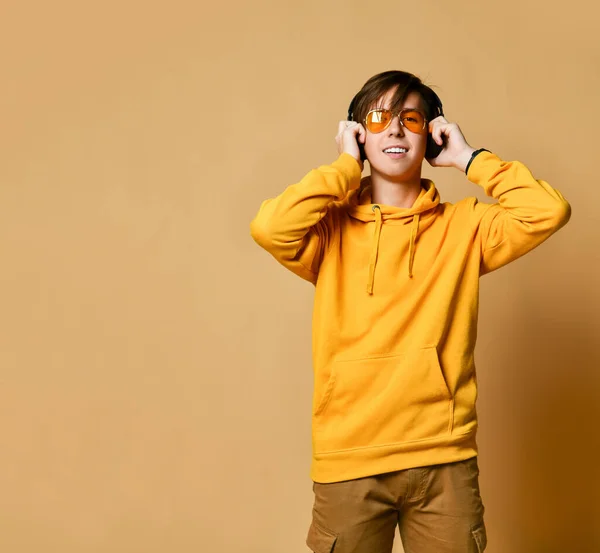 Joven adolescente positivo con capucha amarilla, gafas de sol y auriculares de pie y sonriendo —  Fotos de Stock