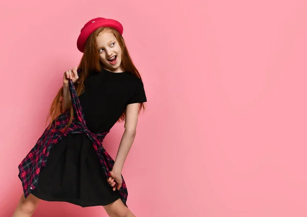 Een tiener in een zwarte jurk en een rode hoed. Ze strakt geruite shirt op haar taille terwijl ze poseert tegen een roze achtergrond. Sluiten. — Stockfoto