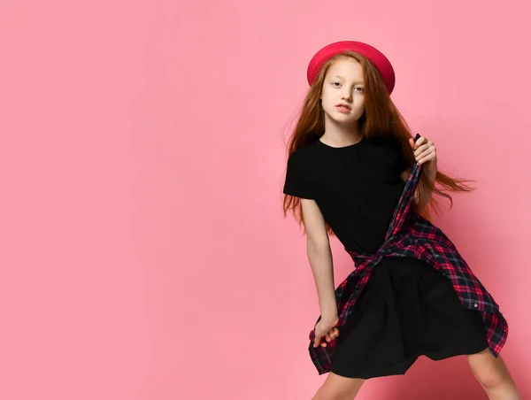 Teenage kid in black dress and red hat. She tightens checkered shirt on her waist while posing against pink background. Close up — ストック写真