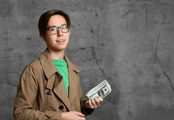 Young teen boy in brown costume and glasses standing and counting dollars over grey concrete wall background