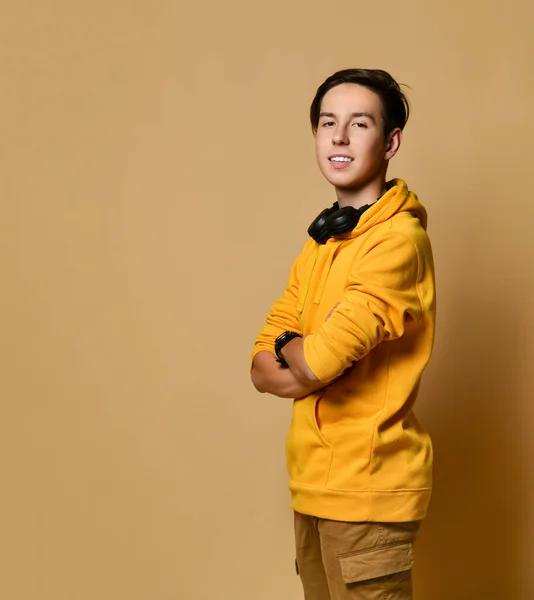 Joven adolescente positivo en sudadera con capucha amarilla y auriculares de pie, sonriendo y mirando a la cámara sobre fondo amarillo de la pared —  Fotos de Stock