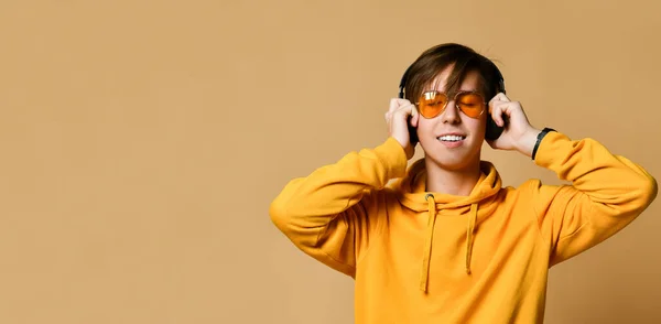 Joven adolescente positivo con capucha amarilla, gafas de sol y auriculares de pie y sonriendo — Foto de Stock