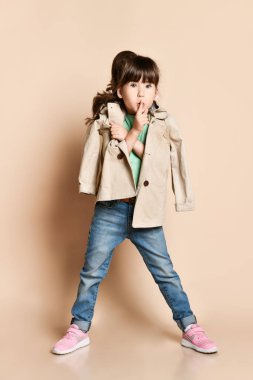 a brunette girl in a beige raincoat makes a hand gesture.