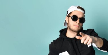 Hipster guy in cap, black tracksuit and sunglasses. He is smiling and showing on you posing on blue background. Close up