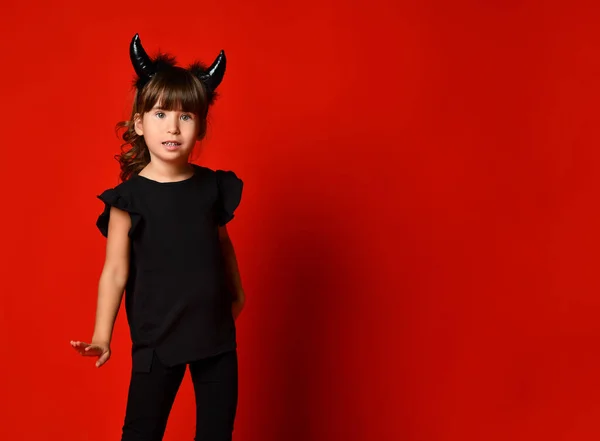 Niña con cuernos de diablo, en blusa negra y polainas posando sobre fondo rojo del estudio. Mascarada, Halloween. De cerca. — Foto de Stock