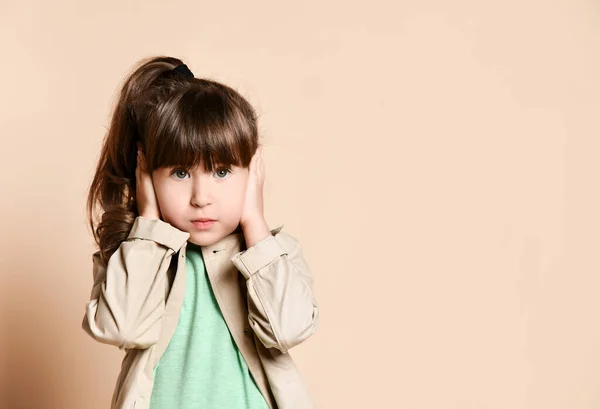 A girl in a beige coat stands with her hands over her ears. — Stockfoto