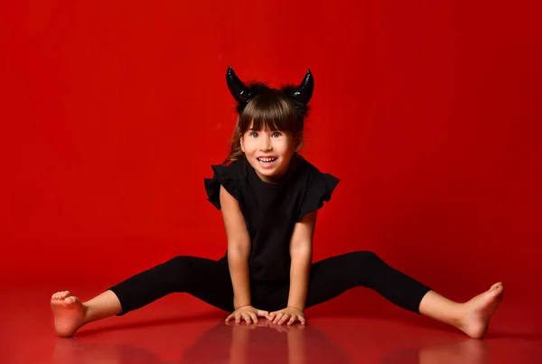 Child with devil horns, in black blouse and leggings. She is smiling, sitting on floor with legs apart. Red background. Close up — Stockfoto