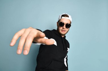 Hipster male in cap, black tracksuit and sunglasses. He is pointing at you by his forefinger, posing on blue background. Close up