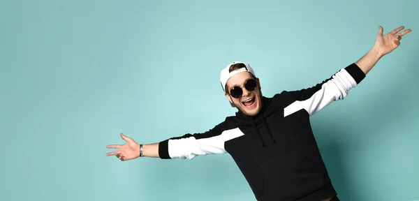 Hipster person in cap, black tracksuit and sunglasses. He spread his arms wide and laughing, posing on blue background. Close up — Φωτογραφία Αρχείου