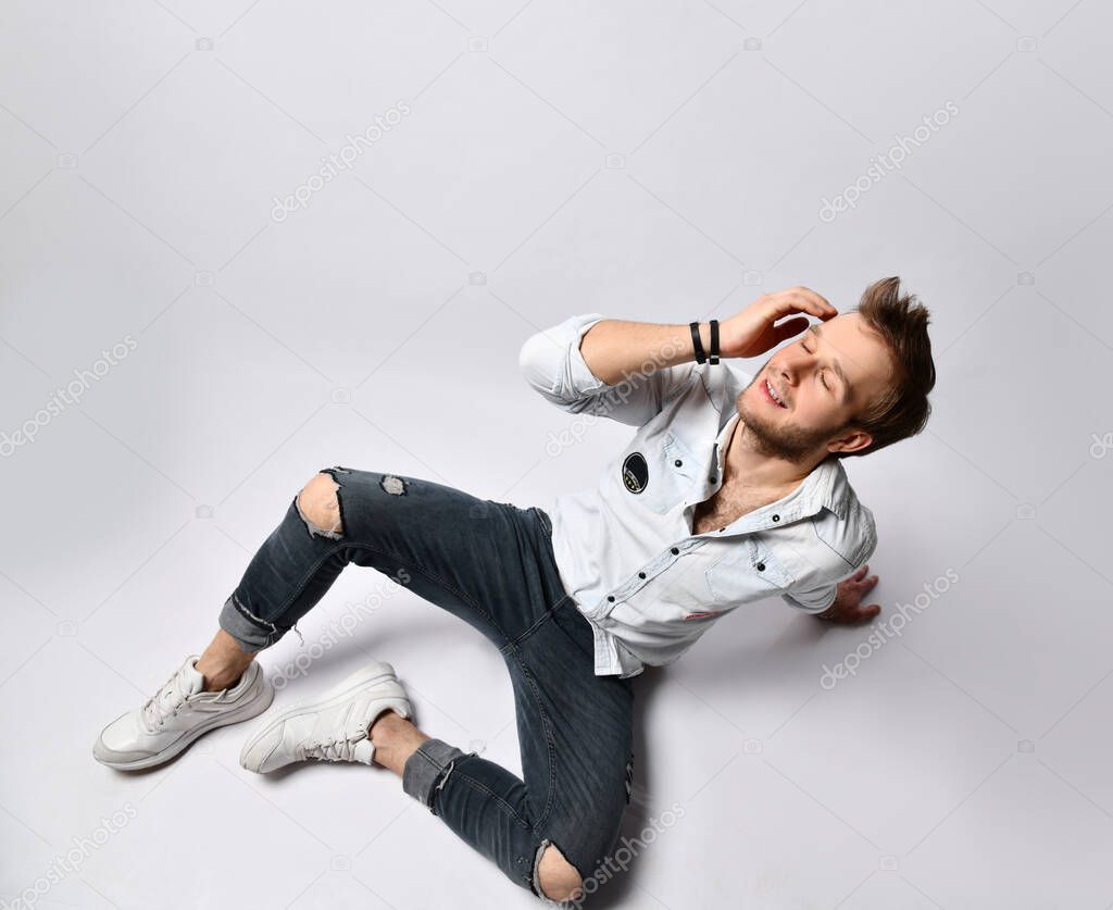Hipster man in shirt, bracelets, jeans and sneakers. He smiling with closed eyes, touching face posing isolated on white. Close up