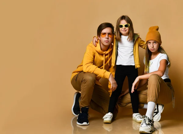 Adolescentes jóvenes y niña pequeña en ropa casual cómoda y gafas de sol sentados y mirando a la cámara sobre el fondo amarillo — Foto de Stock