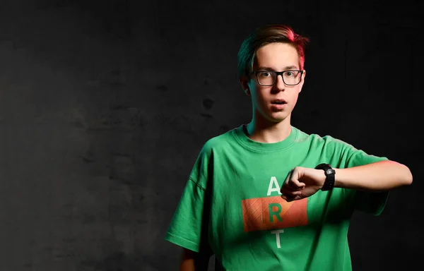 Joven adolescente concentrado en camiseta verde y gafas de pie y mirando el reloj en la mano sobre fondo oscuro —  Fotos de Stock