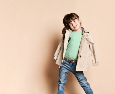 Cute girl posing standing in different poses, in jeans with belt, green t-shirt and beige coat