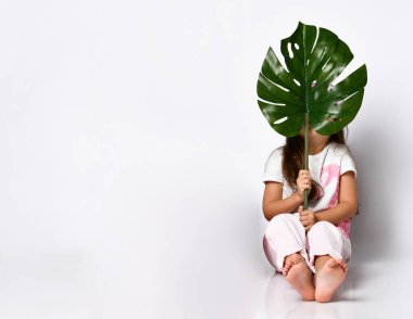 little girl in summer pastel clothes sits on the floor, covering her face with a large crested sheet of tropical raster