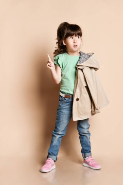 a surprised baby girl stands with a beige coat on her shoulder. Holding your index finger up