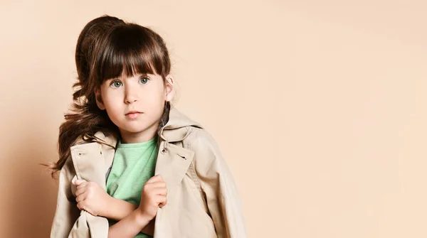 A little brunette girl in a beige raincoat, with her arms crossed over her chest — Stockfoto