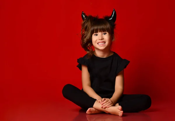Klein meisje met hoorns, in zwarte blouse en legging. Ze glimlacht, zit op de vloer in lotuspositie. Rode achtergrond. Sluiten. — Stockfoto
