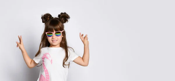 Una pequeña chica alegre, en gafas oscuras en un estudio blanco. Se levanta con las manos en alto y muestra el gesto de paz . — Foto de Stock