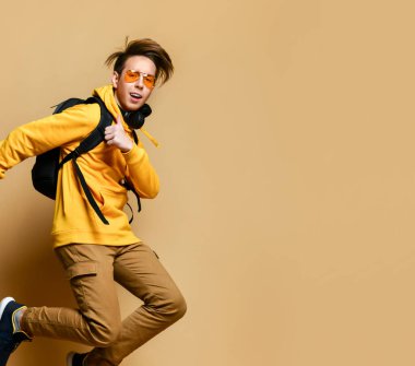 Rahat kıyafetler, spor ayakkabılar, güneş gözlükleri ve sarı bir duvarın üzerinden atlayan sırt çantalı genç bir çocuk.. 