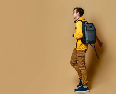 Rahat giyinen, spor ayakkabılı ve sırt çantalı genç bir çocuk sarı duvar arkasında yürüyor..