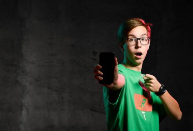 Yeşil tişörtlü ve gözlüklü genç çocuk ayakta duruyor, telefon ekranını gösteriyor ve karanlık arka planda hayal kırıklığına uğruyor.